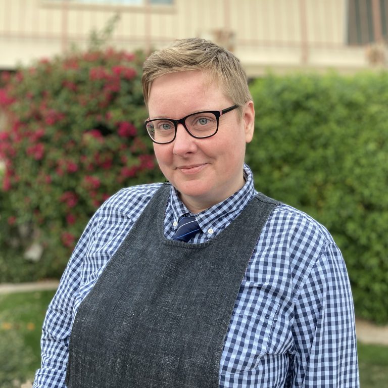 A white, short haired blonde woman with glasses smiles at the camera, wearing a plaid shirt with a tie under a denim pinafore. She stands outside in front of floral bushes and a brick wall.