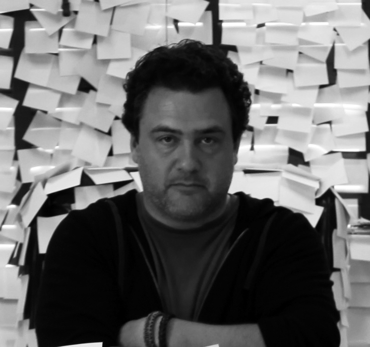Black and White photo of Leopoldo Gout standing with his arms crossed and a wall full of sticky notes behind him