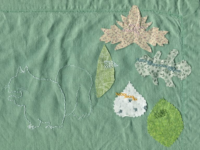 Green linen embroidered with the white outline of a squirrel, and five leaf shapes cut from multiple colors of fabric secured to the linen with cross stitches in various colors.