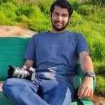 Aravind sits casually on a green bench in park wearing a blue t shirt and holding a camera. He dark hair and a beard and smiles directly at the camera.